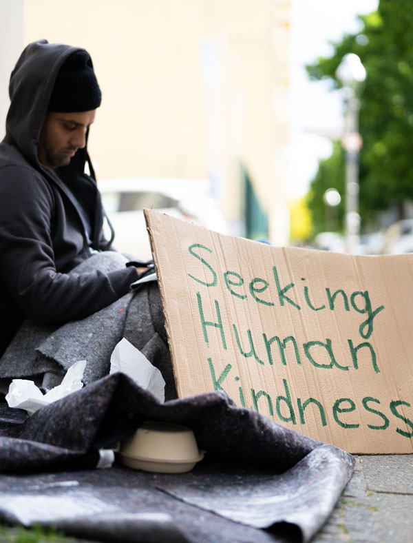 Homeless person on street with cardboard sign that says Seeking Human Kindness